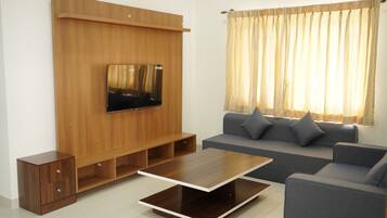 A modern living room with a flat-screen TV mounted on a wooden wall, a grey sofa with cushions, a wooden coffee table, and a window with curtains.