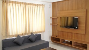A modern living room with a grey sofa, a wooden TV stand, and a window with sheer curtains. - VISTA LIVING (Kasavanahalli)