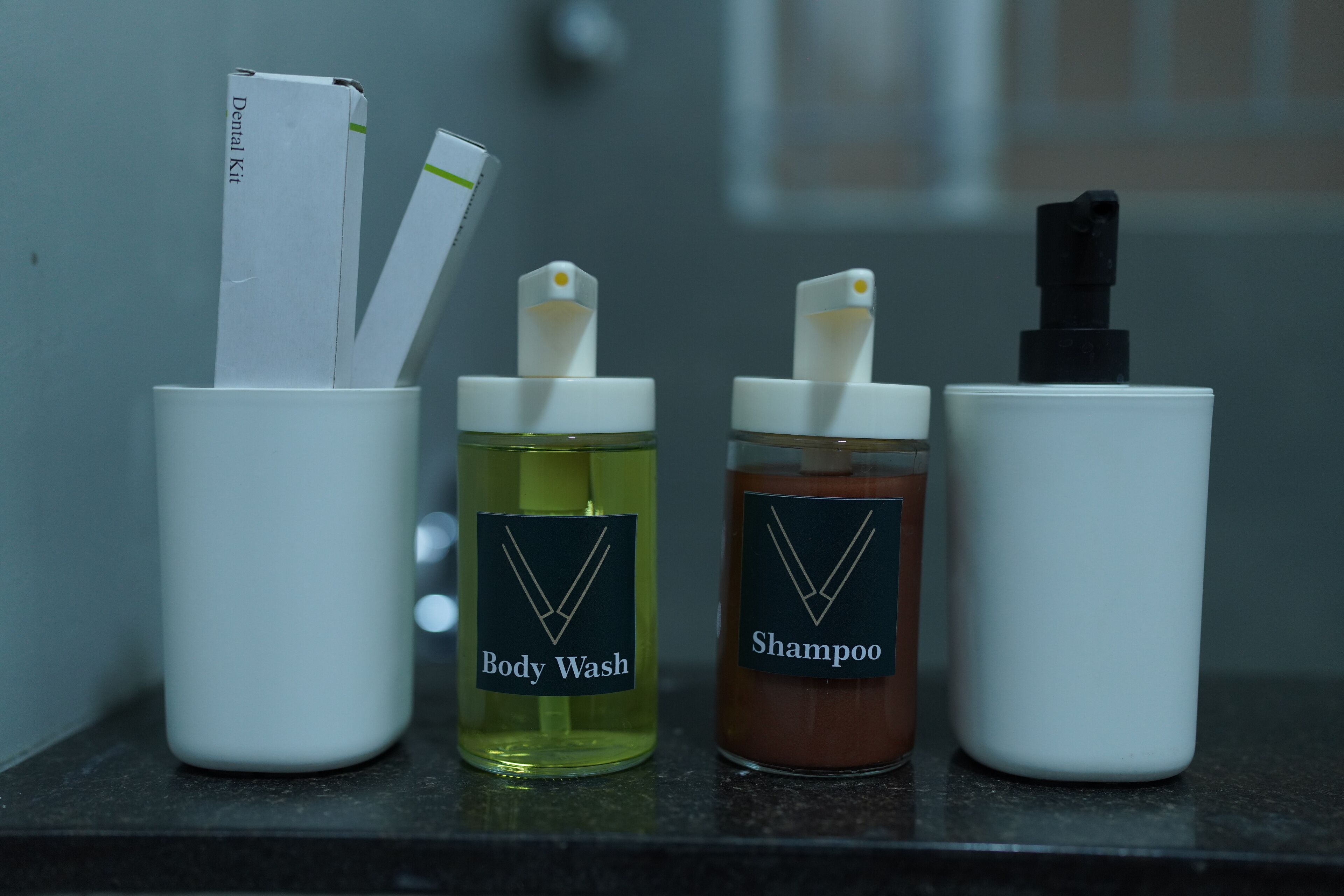 Cuatro recipientes blancos sobre una encimera, cada uno etiquetado con un producto diferente: Gel de ducha, Champú y dos otros con etiquetas que no se ven.