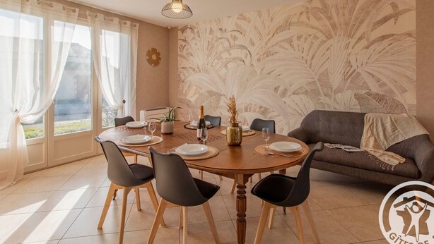 Un comedor con una mesa redonda dispuesta para cuatro personas, rodeada de sillas, un sofá y una pared decorativa con diseño de hojas.