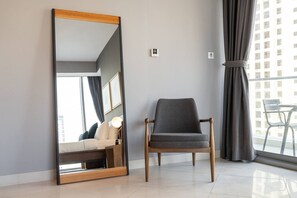 Un dormitorio moderno con un espejo grande, un sillón tapizado en gris, una cama con cabecera blanca y vistas a un paisaje urbano a través de amplios ventanales.