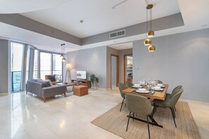 Uma sala de estar moderna com sofá, mesa de centro e televisão, uma área de jantar com mesa posta para seis pessoas e grandes janelas com vista para a cidade.