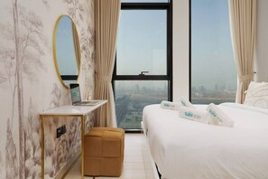 Une chambre d’hôtel avec un lit, un bureau avec un ordinateur portable, un petit pouf, une grande fenêtre donnant sur la ville et un miroir au mur.