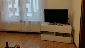 Une pièce dotée d’un téléviseur à écran plat posé sur un meuble blanc, d’un plancher en bois et d’une fenêtre ornée de rideaux légers.