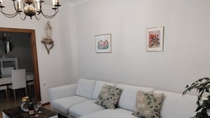 Un salon moderne avec un canapé d’angle, une petite table basse et un lustre. Deux œuvres d’art encadrées ornent le mur, et une plante en pot se trouve dans un coin.