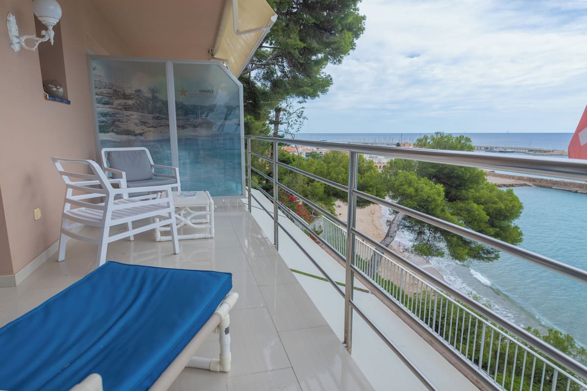 A balcony with a view of the sea, a lounge chair, a sunbed, and a glass door with a mural.