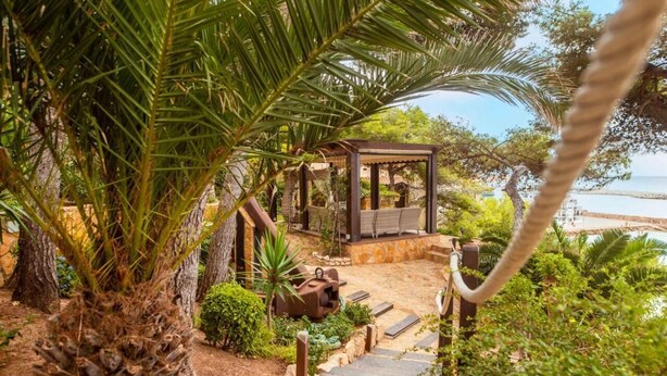 Un espacio de estar al aire libre con techo y vistas al mar, rodeado de abundante vegetación y un sendero de piedra.