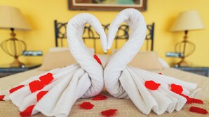 Una cama con una toalla dispuesta en forma de corazón y pétalos de rosa roja sobre una superficie con textura.
