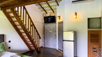 A room with a wooden staircase, a bed with a green bedspread, a refrigerator, and a television mounted on a stone wall.