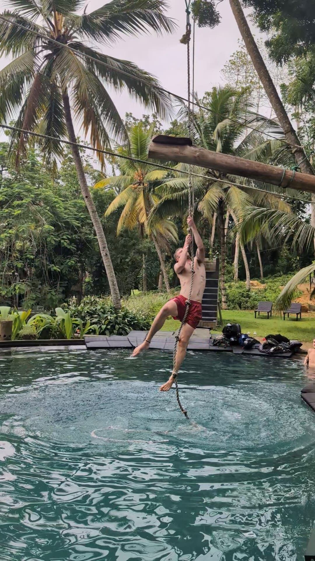 Un hombre colgando de una cuerda sobre una piscina en un entorno tropical con palmeras y una terraza de madera.
