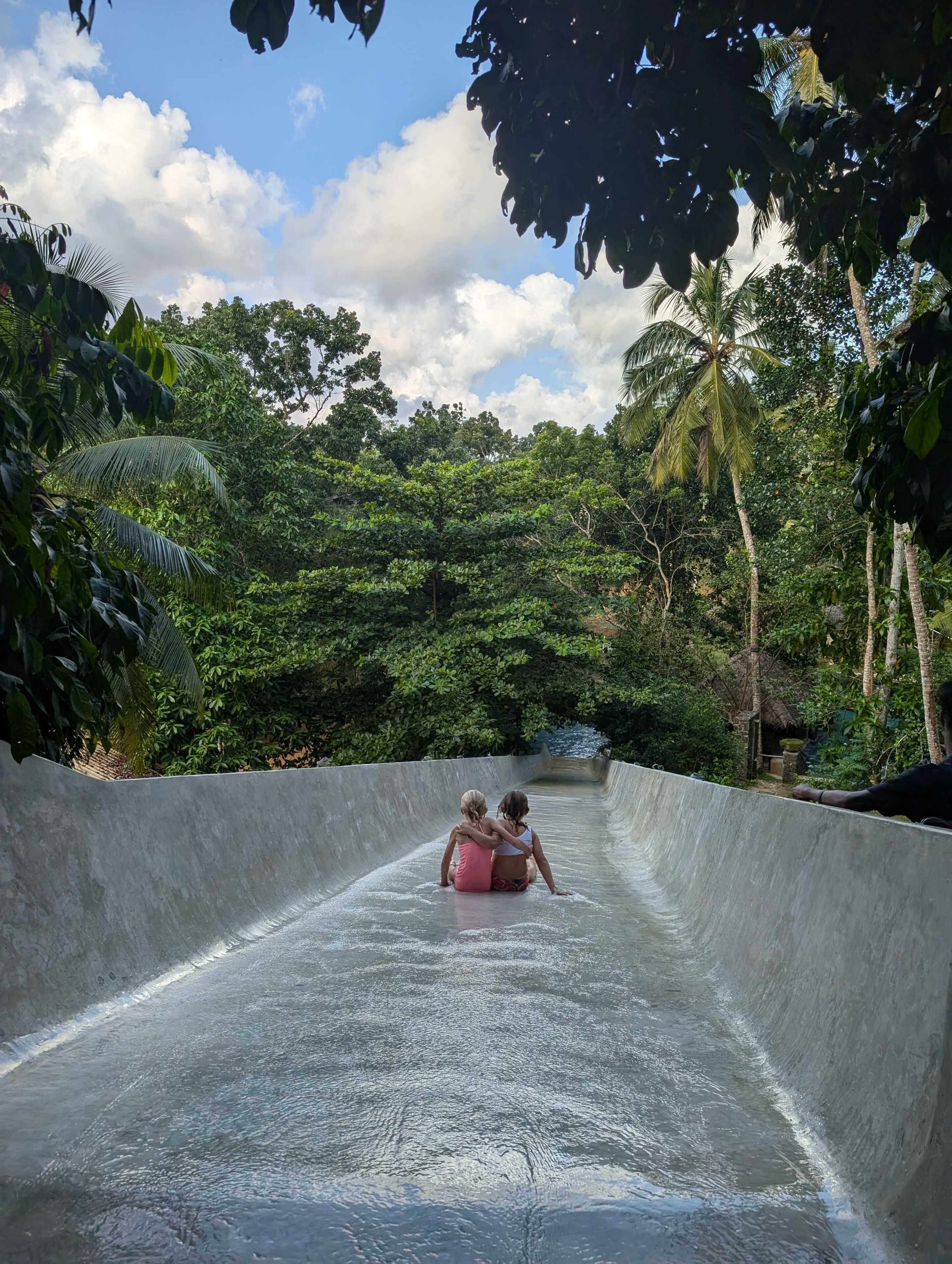 A water slide with two people at the bottom, surrounded by dense tropical foliage.