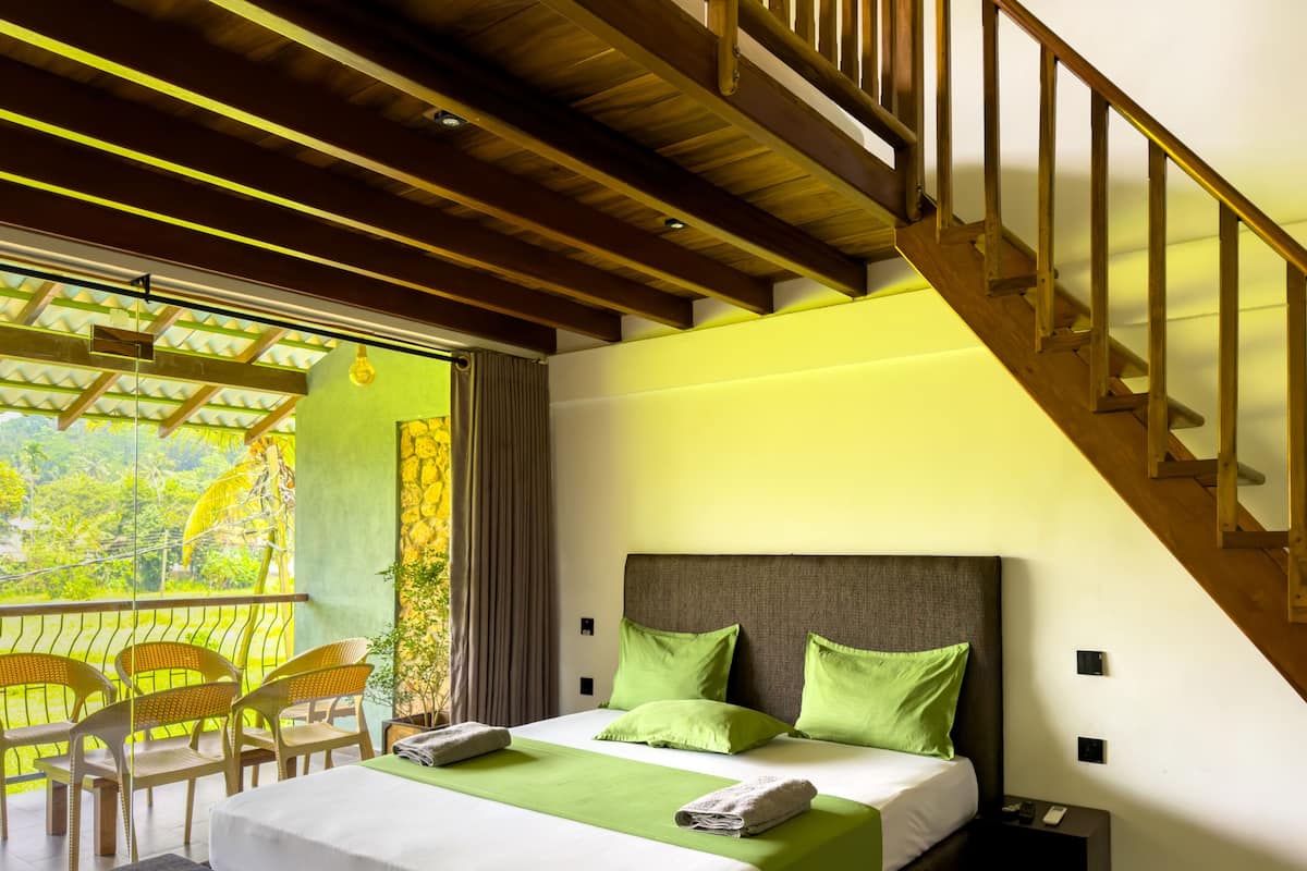 A bedroom with a bed, green pillows, a wooden headboard, a balcony with chairs, and a staircase.