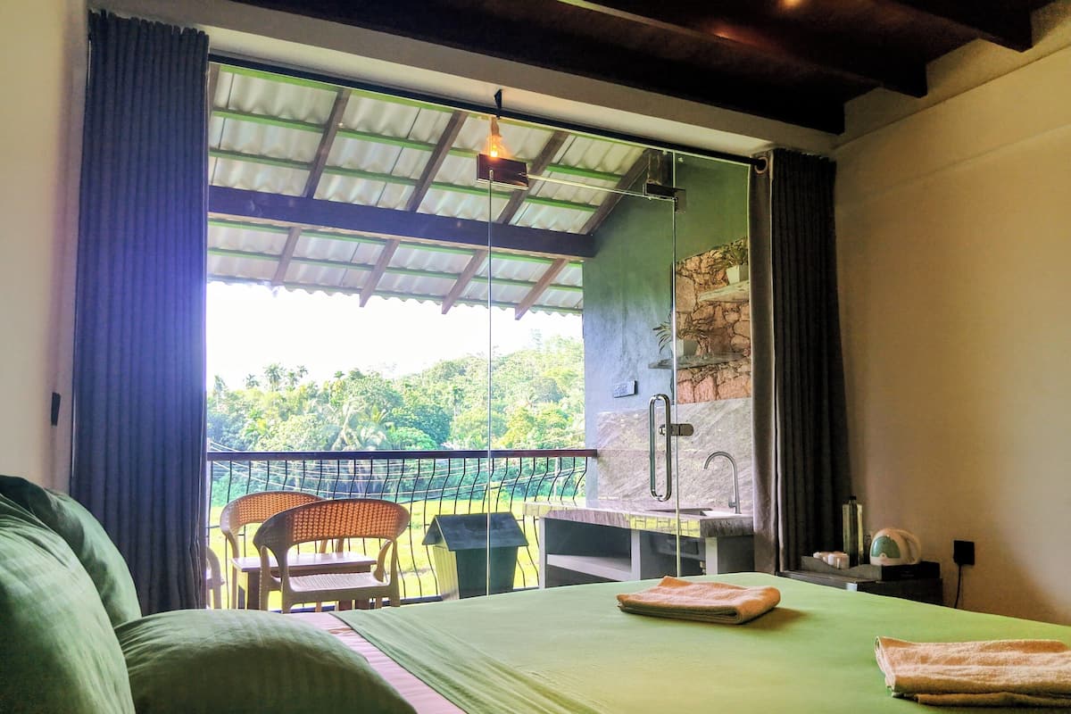 A room with a bed, a small table, a chair, a balcony with a view of greenery, and a shower area.