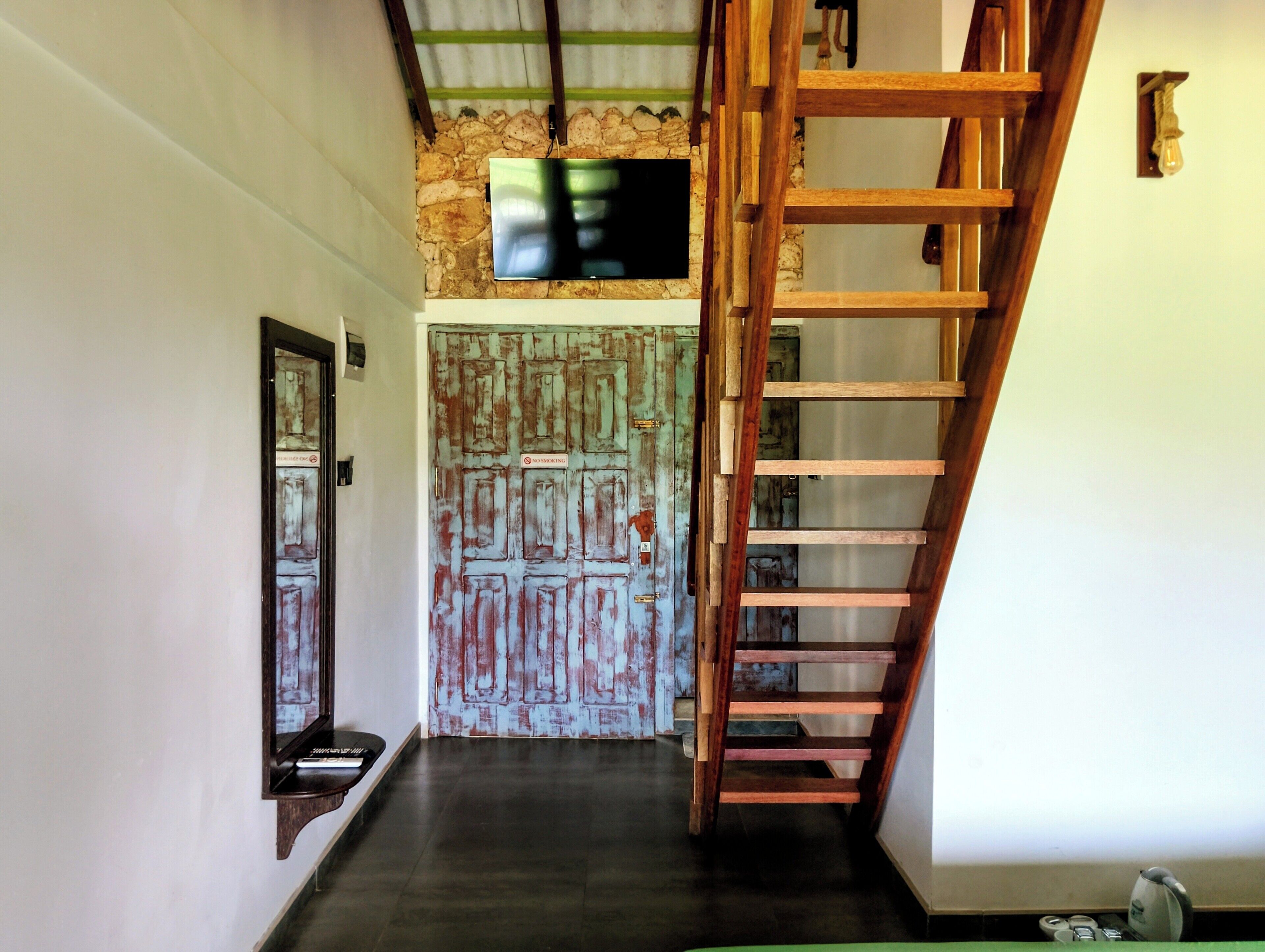 A staircase with a wooden handrail leading to a landing, a stone wall with a television mounted above, and a door with a 'No Smoking' sign.