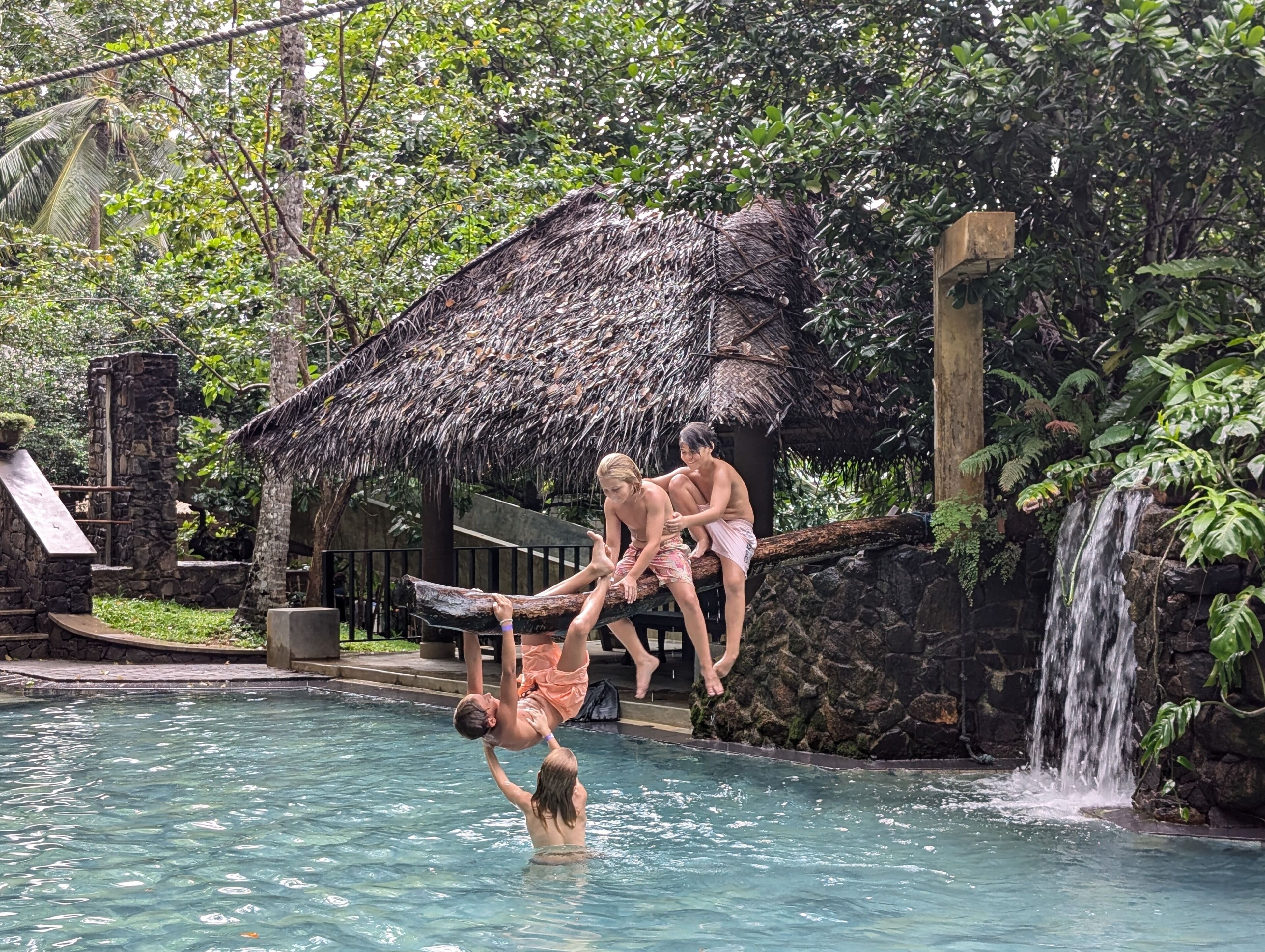 Une piscine avec une cascade, une structure à toit de chaume et un mur en pierre.