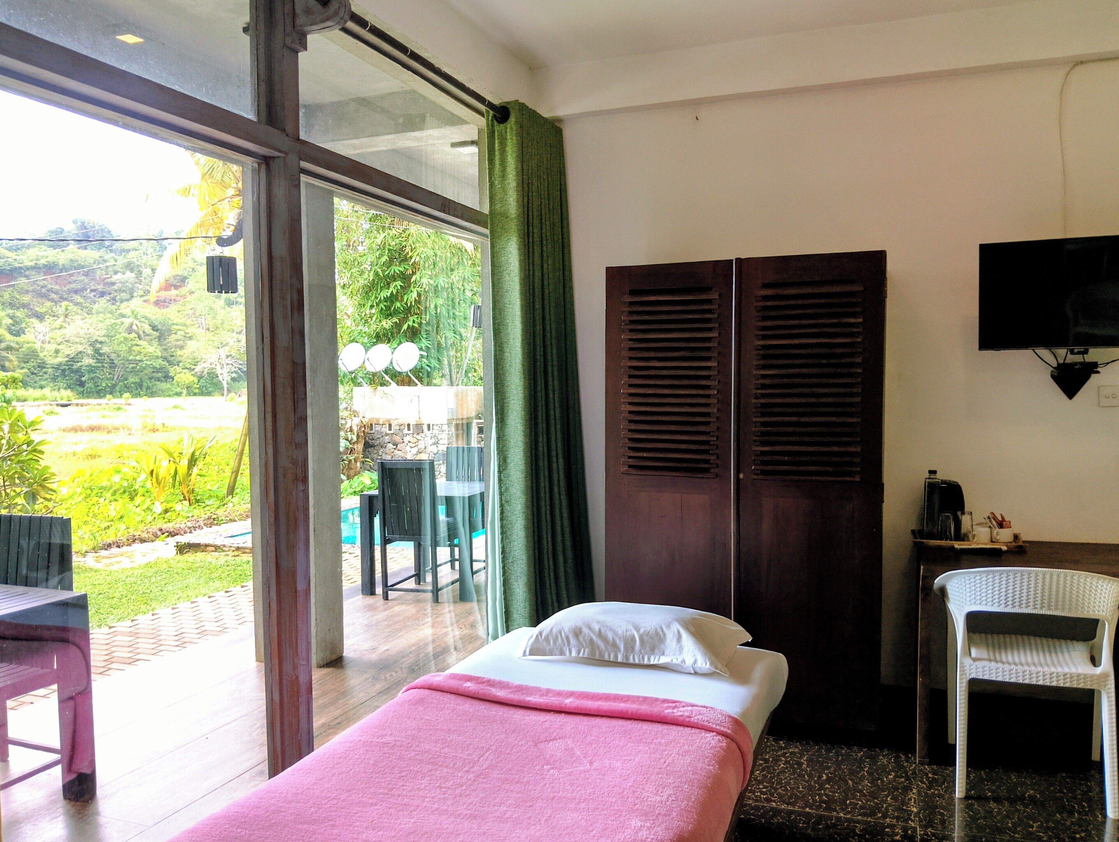 A room with a bed, a TV, a desk with a chair, and a view of a garden through a glass door.