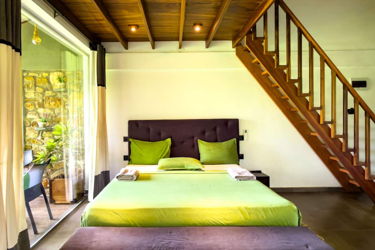 A bedroom with a bed, green pillows, a wooden headboard, a staircase, and a view of a stone wall and plants.