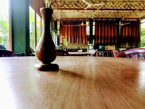 Een houten tafel met een vaas op de voorgrond, een bakstenen plafond en houten gordijnen op de achtergrond.