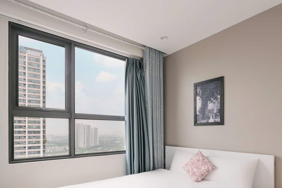 A bedroom with a large bed, a view of tall buildings through a window, and a ceiling fan.