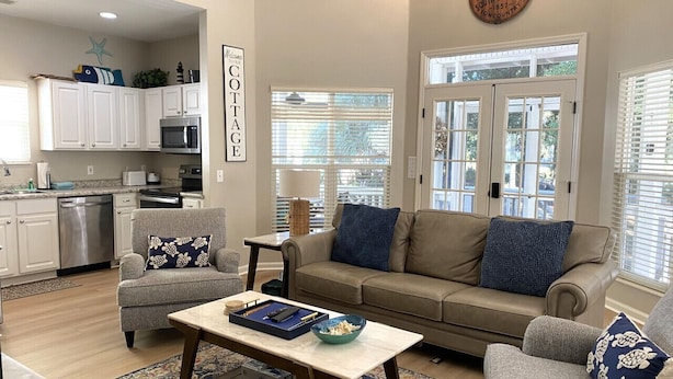 Un salón espacioso con un sofá color beige, dos sillones, una mesita de centro y una zona de cocina con armarios blancos y un microondas.