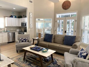 A spacious living room with a beige sofa, two armchairs, a coffee table, and a kitchen area with white cabinets and a microwave. - 26 Davis Love - Cozy Cottage - Owner Cart Incl (Fripp Island)