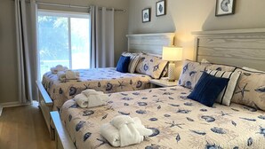 Habitación de hotel con dos camas, cada una con una colcha y almohadas con motivos marinos. Cuenta con una ventana con cortinas, una mesita de noche con lámpara y cuadros enmarcados en la pared.