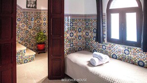 Une pièce avec un mur recouvert de carreaux décoratifs, un lit avec une serviette roulée, une plante en pot et une fenêtre avec un rideau.