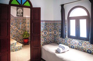 A room with a patterned tile wall, a bed with a rolled towel, a potted plant, and a window with a curtain. - BELLEVUE HOUSE – Entire Home with Panoramic Rooftop in Heart of Medina (Chefchaouen)