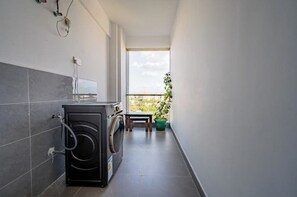 Compact utility area with a washing machine and small table, featuring a tiled floor and a window with a view of the surrounding greenery.