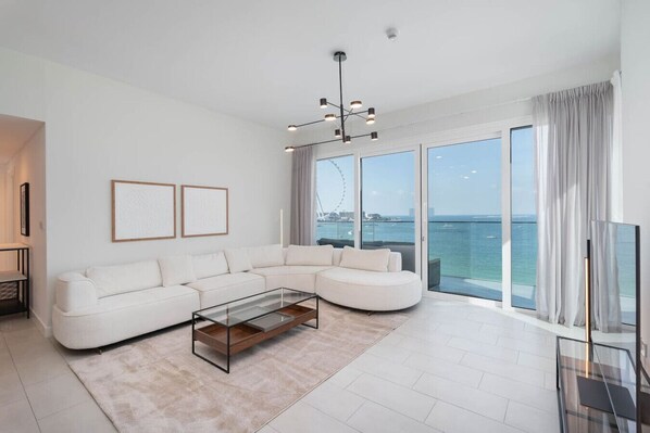 A modern living room with a white sectional sofa, a glass coffee table, and large windows offering a view of the sea and a Ferris wheel.