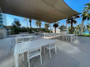 Un espace extérieur aménagé en terrasse, avec des tables et des chaises blanches, un grand auvent et des palmiers.