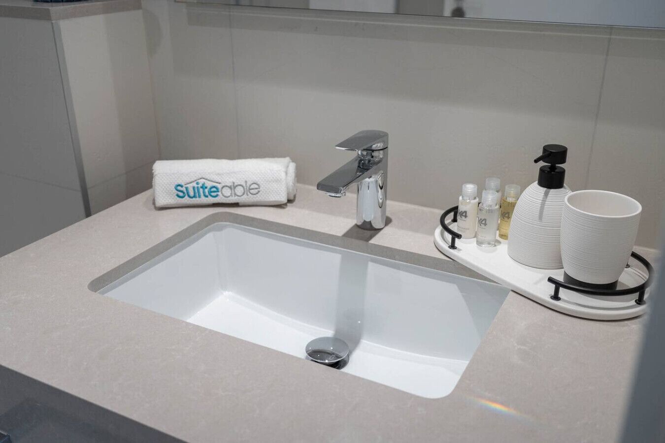 Et moderne badeværelse med en stor vask, et håndklæde med 'Suiteable' branding, en hane og toiletartikler på en bakke.