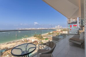 A balcony with a glass railing overlooking a beach and ocean, featuring a round table and two chairs.