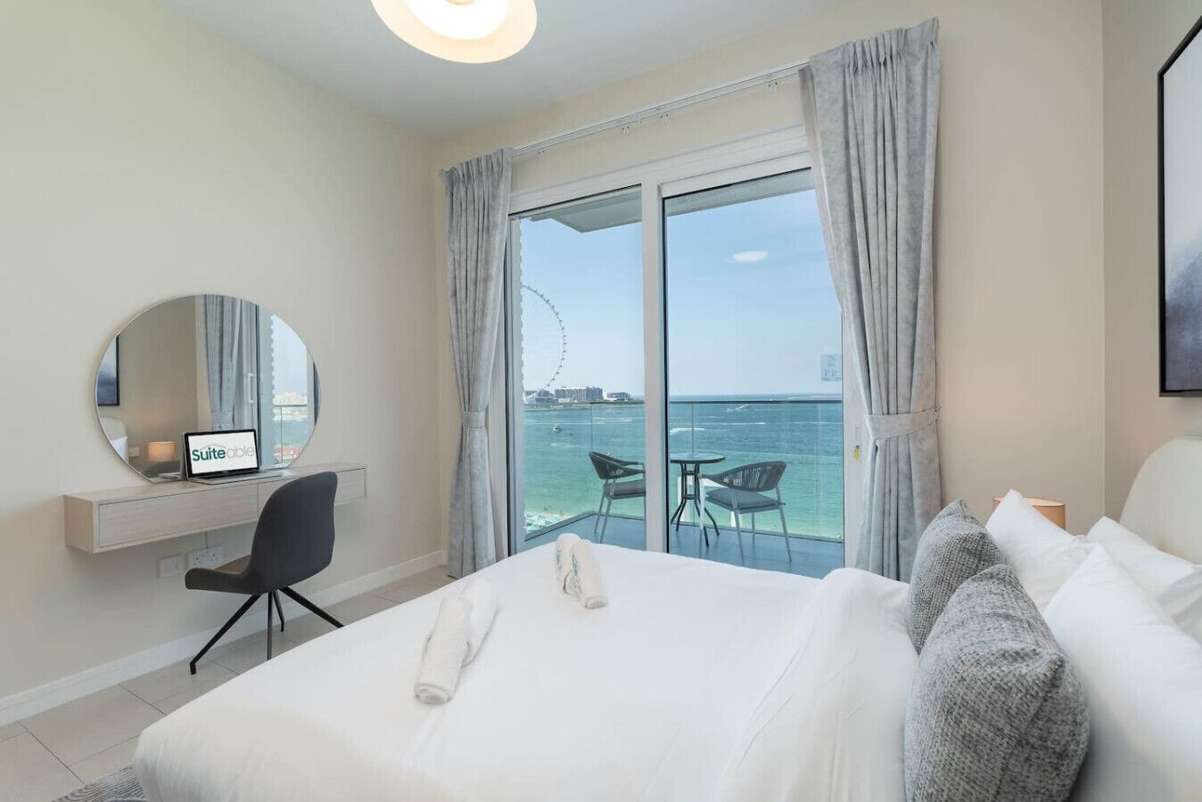 A hotel room with a bed, a desk with a laptop, a chair, and a large window with a view of the sea and a Ferris wheel.