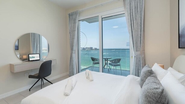 Hotelkamer met een bed, een bureau met een laptop, een stoel en een groot raam met uitzicht op zee en een reuzenrad.