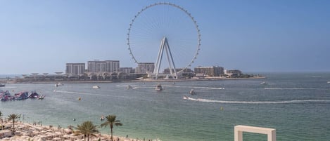 A beach with a large Ferris wheel in the background, a marina with boats, and a sandy area with umbrellas.