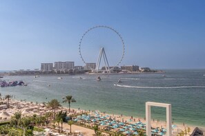 A beach with a large Ferris wheel in the background, a marina with boats, and a sandy area with umbrellas.