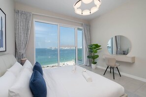 Une chambre d’hôtel avec un grand lit, un bureau, une chaise, une plante en pot, un miroir rond et une vue sur la mer.