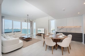 A modern living room with a sofa, dining table, and chairs. There is a television on the wall and a view of the sea through large windows.