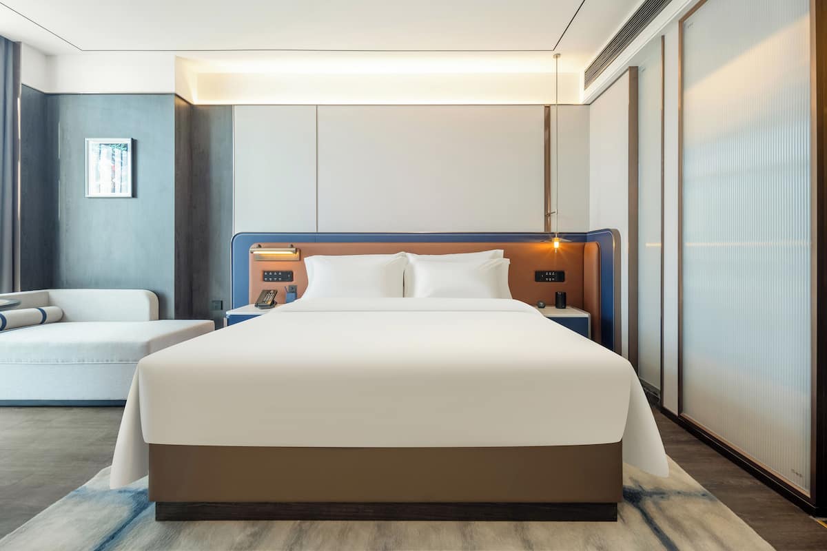 Una habitación de hotel moderna con una cama grande, una mesita de noche con lámpara, una pequeña zona de estar con sofá y un ventanal con persianas.
