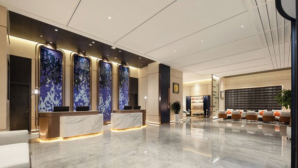 Un hall d’hôtel moderne avec un comptoir d’accueil en marbre, un espace salon doté de coussins orange et un grand mur orné d’un motif géométrique.