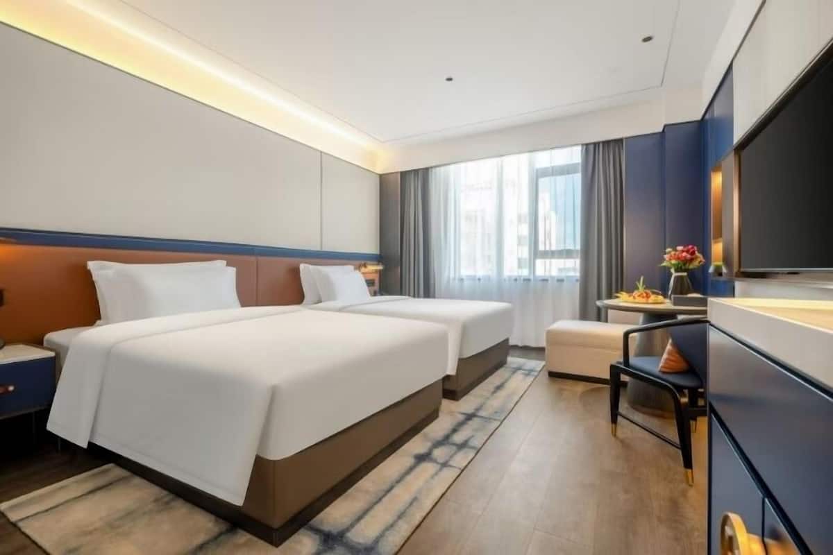 Habitación de hotel con dos camas, un televisor de pantalla plana, una mesita con un jarrón de flores y vistas a la ciudad a través de amplios ventanales.