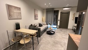 A modern living room with a sofa, coffee table, and dining area. There is a door leading to the kitchen, and a TV mounted on the wall. - Meliana Homes (Parga)