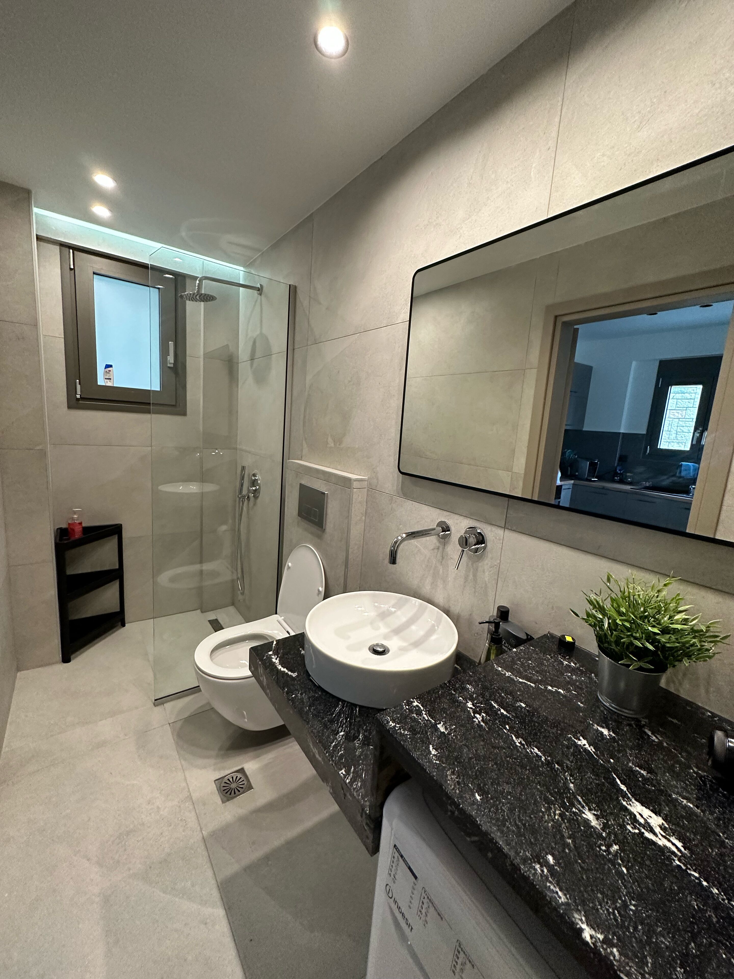 Une salle de bain moderne avec une douche à parois de verre, des toilettes blanches, un lavabo rond doté d’un plan de travail en marbre et un grand miroir.
