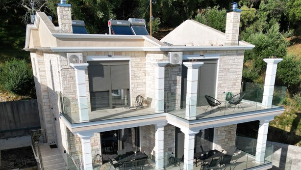 A modern two-story house with a glass balcony, outdoor seating area, and a stone facade. - Meliana Homes (Parga)