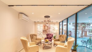 A modern living room with beige armchairs, a round coffee table with flowers, and a large window with a view of the street.