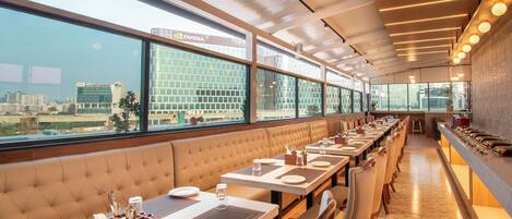 A modern restaurant with long tables, cushioned booths, and large windows offering city views.