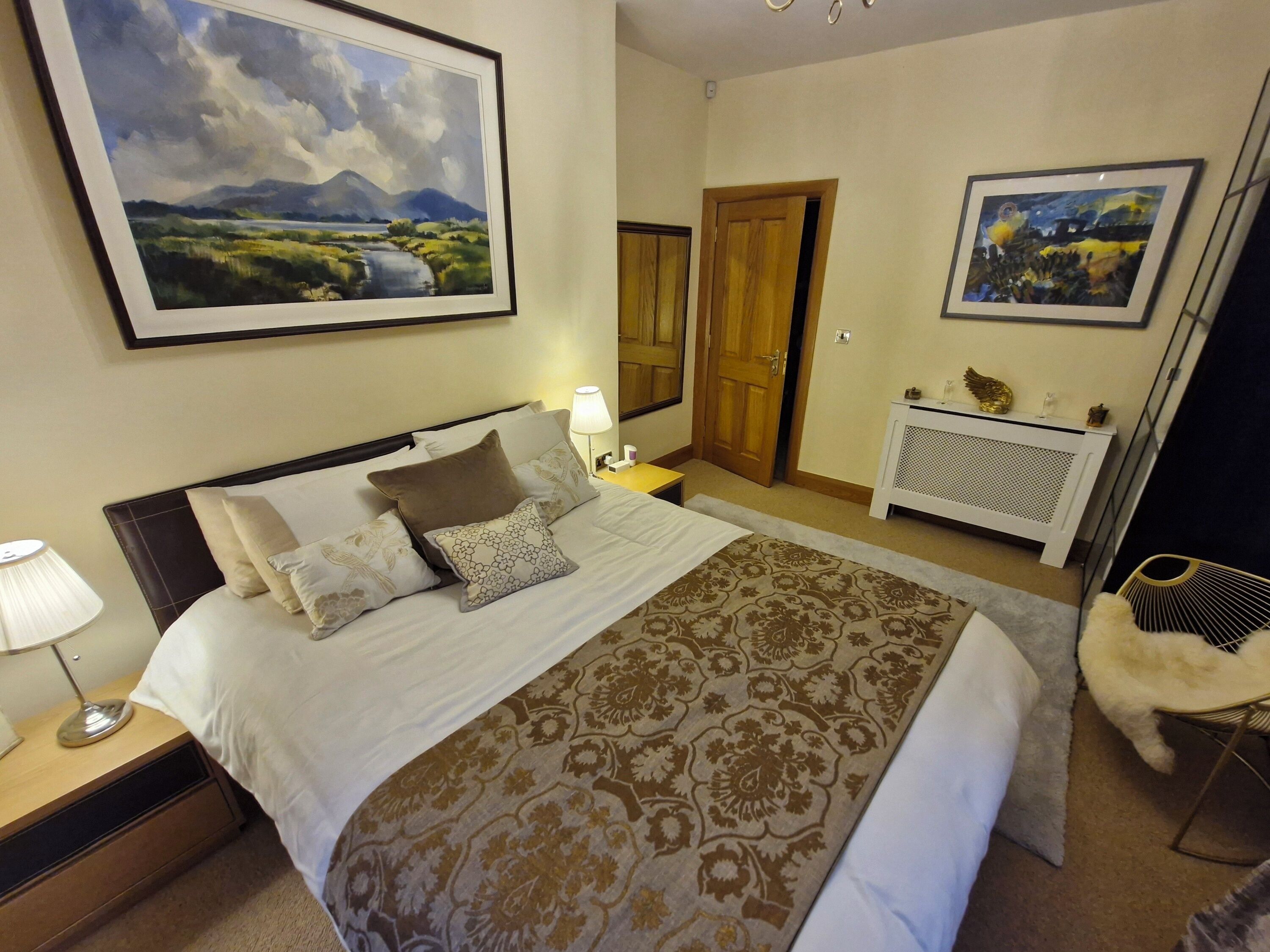 A neatly made bed with a patterned bedspread, a bedside table with a lamp, a framed landscape painting above the bed, and a radiator with a decorative item on top.