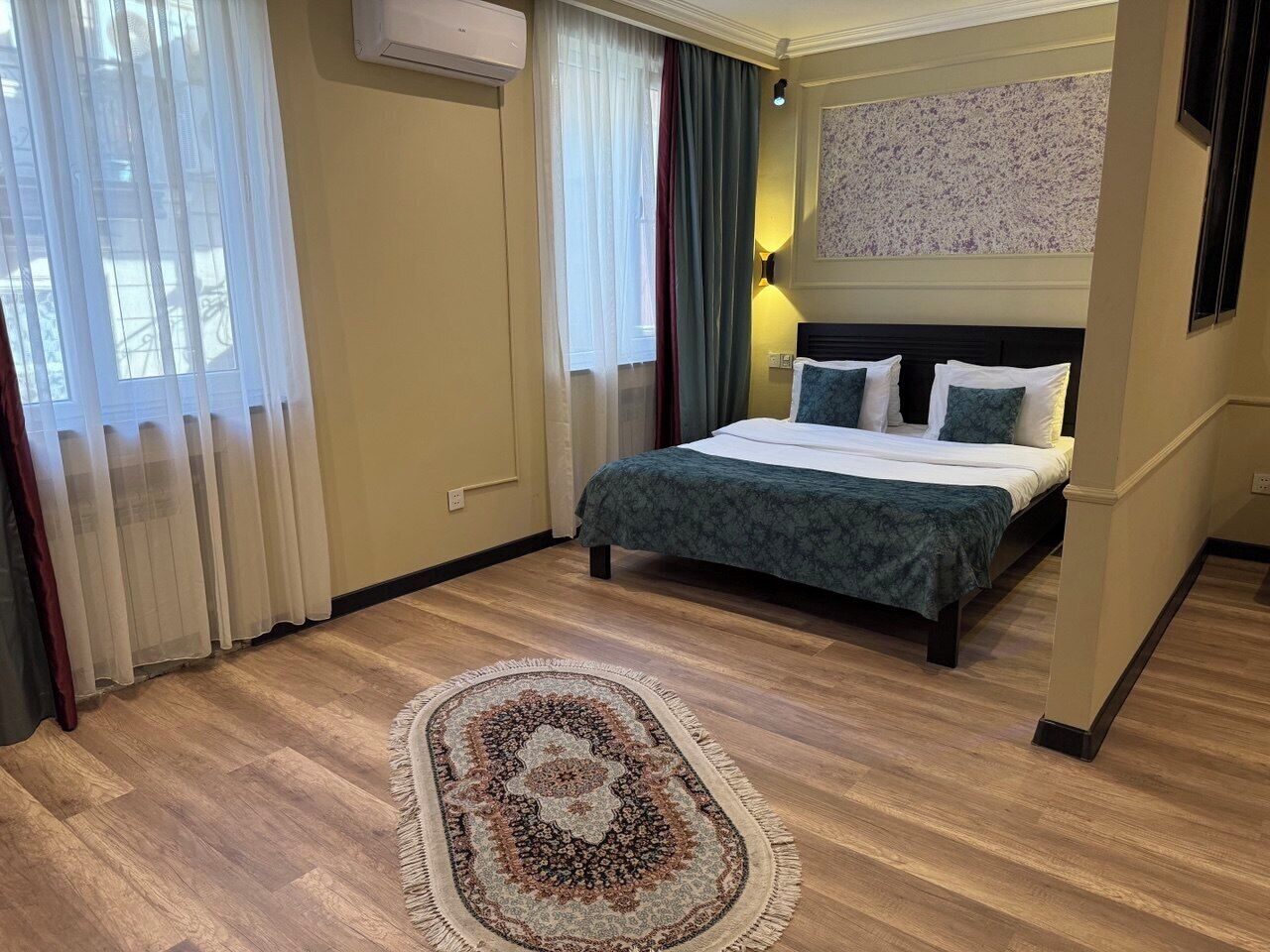 A hotel room with a bed, a patterned rug, curtains, and a wall-mounted air conditioner.