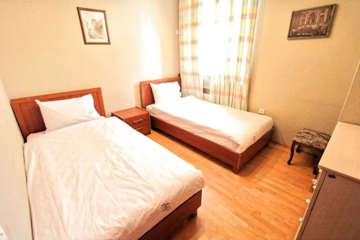 A hotel room with two single beds, a wooden nightstand, a small table, and a bench.